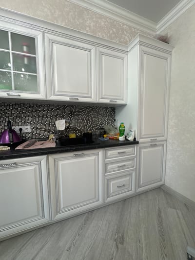 The image displays a high-angle view of a modern kitchen interior in Yuzhny, Russia, rotated 90 degrees clockwise. The kitchen features an L-shaped layout with light gray or off-white cabinetry, extending to the ceiling. The cabinet doors are adorned with ornate raised panels and decorative silver trim, incorporating a Greek key pattern, complemented by silver-toned handles. One of the lower cabinets on the left side of the frame has a glass-paneled door.

The walls are covered with a light, textured wallpaper, bordered by white crown molding at the ceiling. The floor, visible on the right, features light gray, wood-look laminate or tile planks.

A dark countertop runs along the base of the cabinets. A striking black and white mosaic tile backsplash fills the area between the upper and lower cabinets, particularly visible around the sink section. On the countertop, several items are arranged, including a green bottle labeled "АКТИБИО", various plastic bags, a sponge, and a pinkish cloth. A white electrical outlet with multiple plugs is situated above the countertop, connected to a purple small appliance, possibly a blender or kettle.

No people are visible in the scene, and no specific activity is occurring, presenting a static snapshot of the kitchen space. The lighting indicates it is daytime.