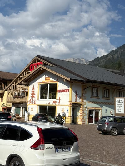 The image shows a building with a Coop Market sign on its facade, located in Campitello di Fassa, Italy. The building has a distinctive alpine architectural style with wooden balconies and decorative paintings. In front of the building, there is a paved parking area with several cars and a motorcycle with a rider. The sky is partly cloudy, and a mountain range is visible in the background. The Coop Market appears to be a local grocery store or supermarket. A signpost to the right of the building lists "Tobia" with services like "Restaurant, Wine Bar, Pizza, Caffetteria, Garai."