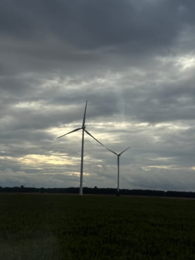 A pair of wind turbines stand silhouetted against a dramatic, overcast sky in a rural landscape. The sky is filled with dark, heavy clouds with patches of lighter sky peeking through near the horizon, suggesting either dawn or dusk. The left turbine, closer to the viewer, is larger and more in focus, with its three blades spread wide. The second turbine, smaller and further in the distance, is also visible. Both turbines are positioned in a vast, flat field of dark green vegetation, possibly crops. A dark line of trees forms a distant horizon behind the field. The scene evokes a sense of open space and a moody atmosphere due to the weather conditions. The location is the city of Sieradz, Poland.