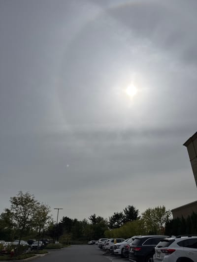 The image displays an overcast sky over a commercial parking lot in Plainsboro Township, United States. A bright, sun-like object with a distinct starburst effect is prominently featured in the upper central portion of the frame, surrounded by a faint, partial solar halo visible in the upper left quadrant. A smaller, diffuse luminous spot is present lower and to the left of the main sun. The sky is uniformly gray with thin cloud cover.

The lower half of the image depicts an asphalt parking lot populated by multiple parked vehicles, primarily SUVs and sedans, in various colors including white, black, silver, and dark gray. On the left side, a cluster of deciduous trees with green and some autumnal foliage is visible, along with a dark-colored utility pole. The right side shows a portion of a light-colored building exterior with a visible dark roofline, partially obscured by a dense row of dark green evergreen shrubs. No individuals or specific activities are discernible within the scene. The precise location within Plainsboro Township cannot be determined from the visual information provided.