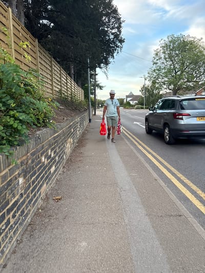 A man wearing a white hat, a light blue t-shirt with the word "HOLLISTER" printed on it, and olive green shorts is walking on a sidewalk. He is carrying two red shopping bags, one in each hand. The man is walking away from the camera towards a road with a grey SUV. The sidewalk is next to a brick wall and a wooden fence. The sky is cloudy with hints of blue. This scene appears to be located on a suburban street in Chelmsford, United Kingdom, possibly near a residential area with trees and houses visible in the background.