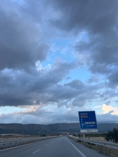 The image depicts a roadside scene on a highway approaching Manfredonia, Italy. The foreground is dominated by the asphalt road, divided by white lines, and a metal guardrail running along the right side. A blue directional sign is prominently displayed on the right, indicating "VIESTE" with an arrow pointing upwards and "Gargano" below. Another directional arrow points to the right for "MANFREDONIA" and "S. GIOVANNI R." below, with "SP 58" visible. A distance marker reads "700 m".

In the background, rolling hills and mountains form the horizon. The sky is filled with dramatic, dark gray clouds with patches of blue sky peeking through. The overall lighting suggests it might be late afternoon or early evening, possibly before or after a rain shower. A few vehicles are visible on the road in the distance. The environment appears to be a rural or semi-rural highway setting.