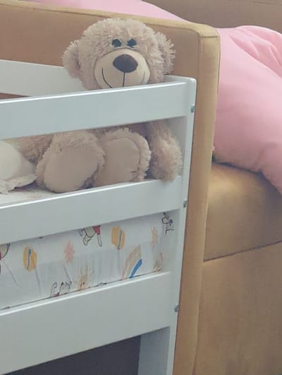 A fluffy, light brown teddy bear rests its head over the top railing of a white crib. The bear's face, with black button eyes and a stitched smile, is visible. Its body is mostly inside the crib, with its paws resting on the mattress. The crib has a white frame with visible horizontal slats. Beneath the bear, the crib mattress is covered with a white sheet adorned with a cartoon pattern featuring Winnie the Pooh and Tigger. The crib is positioned next to a large, light brown upholstered piece of furniture, possibly a sofa or chair, with a bright pink cushion partially visible in the background. The setting appears to be an indoor domestic environment, such as a nursery or bedroom. No specific location in Tbilisi, Georgia can be determined from the image.