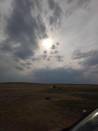 The image shows a wide, open, flat landscape under a dramatic, cloudy sky. The sun is partially visible through the clouds, creating a diffused light. The ground is covered in dry, yellowish-brown grass, suggesting a steppe or prairie environment. In the middle ground, there are some scattered dark objects that appear to be discarded tires or similar debris. The foreground shows a portion of a dark-colored vehicle, likely a car, with its windshield reflecting the sky. The scene is devoid of any people or animals. The overall impression is one of a remote, desolate, and possibly windy rural area, consistent with the description of the city of Sartymbet, Kazakhstan, which is known for its steppe landscape.