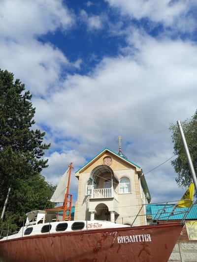 In Giaginskaya, Russia, a peculiar scene unfolds featuring a large, rust-colored boat with white accents situated in front of a light-colored building resembling a church with a blue roof and golden cross. The boat has the word "ВИЗАНТИЯ" (Byzantium) written in white lettering on its side. A white sail is visible on a mast at the front of the boat. The building has arched windows and a balcony. Trees flank the scene, and a bright blue sky with scattered white clouds serves as the backdrop. The overall impression is of an unusual, possibly artistic, display.