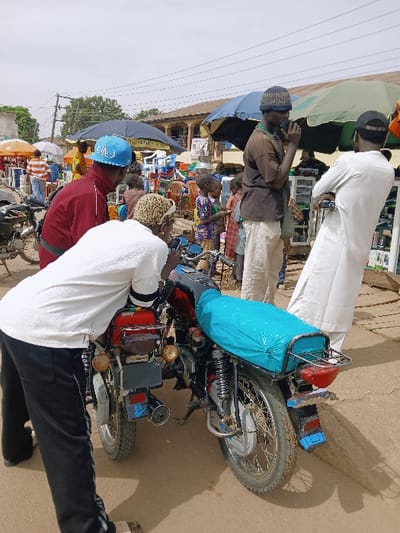 This image captures a busy outdoor street or market scene in Kwekwe, Zimbabwe, under a bright, diffused sky. In the foreground, four adult males are prominent around two motorcycles parked on a light brown, possibly dirt, surface.
On the left, a man wearing a white short-sleeved shirt and black track pants with white side stripes is bent over, facing towards the rear of a motorcycle with a red fuel tank, suggesting inspection or repair. To his immediate left, another man in a red long-sleeved jacket and a blue baseball cap looks down towards the bent man and the motorcycles.
In the mid-ground, on the right, a third man wearing a brown short-sleeved shirt and a dark knit cap stands with a dark object held to his mouth. Adjacent to him, a fourth man, dressed in a long white garment and a dark baseball cap worn backward, stands slightly reclined, holding what appears to be handlebars, facing the group and motorcycles.
The motorcycle in the right mid-ground has a red fuel tank, a black seat partially covered by a bright teal fabric, a rear cargo rack, a red taillight, and a blue and white license plate visible on its mud-splattered rear fender.
The background reveals a bustling market environment with numerous individuals, including children, moving among various stalls. These stalls are sheltered by large, colorful umbrellas in shades of dark blue, green, and orange. Merchandise such as plastic chairs and other household items is visible. Buildings with brown roofs and utility poles with overhead wires line the horizon.