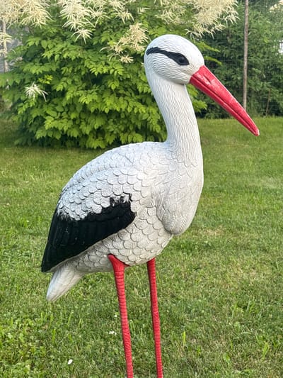 The image shows a decorative stork statue standing on a green lawn. The statue is white with black wings and a red beak and legs. It is positioned in the foreground, with a lush green bush and white flowers in the background. The lighting suggests it is daytime, likely outdoors in a garden or park setting. There is no indication of this location being in the city of Yermolino, Russia.