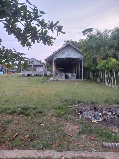 The image depicts an outdoor scene in Gampongkuala, Indonesia, featuring two unfinished or abandoned concrete structures with exposed frameworks and roofs. The structure on the right is open on its sides, supported by pillars, and contains concrete platforms inside. The structure on the left is also supported by pillars and has partially completed walls. Both buildings appear weathered, with peeling paint and visible damage.
The foreground is a grassy area with scattered dry leaves and some litter, including plastic bottles and wrappers. To the right in the foreground, there is evidence of a recent bonfire, with ashes and burnt debris.
In the background, behind the structures, are dense trees, including palm trees and other foliage. A line of slender trees with light-colored trunks stands between the structures and the denser vegetation. Further in the distance, to the left of the structures, a few other buildings with tiled roofs are visible, suggesting a populated area nearby. The sky above is overcast with soft clouds. The overall impression is one of a rural or semi-developed area, possibly a beachside location given the palm trees, with ongoing or halted construction and signs of human activity.