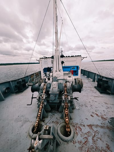 The image is a forward-facing view from the deck of a ship on a body of water, likely a river or lake, with a cloudy sky overhead. In the foreground, there are ship mechanisms, including a winch with a heavy chain extending downwards into a circular opening in the deck. The deck is grey and shows signs of wear and tear, with patches of rust and scuff marks. There are bollards on either side of the deck. In the mid-ground, there is a white structure with blue accents, possibly a cabin or equipment housing, with a ladder leading up to it. The water is calm and reflects the overcast sky. On the horizon, there is a faint green line of trees, indicating land. The image appears to have been taken in Zotino, Russia, though no specific landmarks or urban features are visible to confirm this. There are no people present in the image.