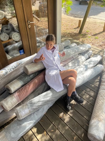 A young woman in a white jacket lies atop a pile of rolled rugs, seemingly on a porch or deck, in an outdoor setting. The rugs are wrapped in plastic and vary in color and pattern. To her left is a window display of more rolled rugs inside a building. Her legs are crossed near the top of the pile, and she looks directly at the camera. Sunlight streams across the scene.
