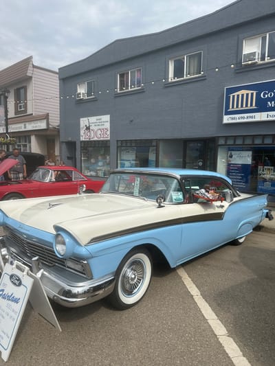 This image captures a classic car show event in Leduc, Canada, likely on a cloudy day. The primary focus is a two-tone blue and white 1957 Ford Fairlane 500 2-Door Hardtop, prominently displayed on a street. The car is in pristine condition, featuring whitewall tires and gleaming chrome accents. A sign in front of the car provides details about its specifications, including engine size, horsepower, and color.
In the background, a row of commercial buildings lines the street. One building, painted gray, features signs for "Dominion Lending Centres," "The Edge" (offering dance, gymnastics, and figure skating), and "Clean My Air." Another building to the right is identified as a restaurant with a "Buffet Takeout Delivery" service. The buildings suggest a downtown or main street setting.
Several people are visible, mostly near the cars, indicating they are attendees or participants in the event. One person is leaning over a red car parked next to the Ford Fairlane, and others are seen walking around. The overall atmosphere appears to be casual and community-oriented, typical of a local car show. The presence of street parking with painted lines and the building facades suggest an urban or town setting. No specific time of day can be definitively determined, but the lighting suggests daytime.