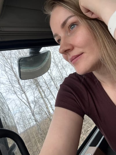 A woman with blonde hair, wearing a burgundy long-sleeved shirt and a white wrist device, is seated inside a vehicle. The vehicle's side mirror is visible, reflecting a view of trees with bare branches, likely a winter or early spring scene. The location cannot be determined as Kukui, Russia based on the image content alone. The steering wheel of the vehicle is partially visible in the lower right corner.