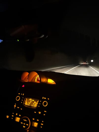 The scene is a nighttime view from the interior of a vehicle traveling on a two-lane paved road in Kalnovo, Bulgaria. The dashboard is visible in the foreground, illuminated with amber-colored light from the gauges (including speedometer and tachometer) and the center console controls for climate and audio systems. A faint, blurred outline of a hand or arm is discernible in the upper left portion of the image, suggesting the presence of a person, likely the driver or a passenger. The vehicle's headlights illuminate the road ahead, revealing solid white lines on the shoulders and a dashed white line marking the center. In the distance, multiple red taillights indicate at least one other vehicle traveling in the same direction, and a circular, reflective sign is visible on the right side of the road. The environment surrounding the road is dark, devoid of visible structures, suggesting a rural or unlit area.