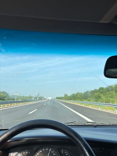 The image depicts a first-person perspective from the driver's seat of a vehicle traveling on a multi-lane highway in the vicinity of Dolni Bogrov, Bulgaria. The road surface is asphalt, with clear white lane markings visible, including a solid white line on the left and a dashed line on the right, indicating movement in the rightmost lane. Metal guardrails line both sides of the highway, beyond which extensive green vegetation, primarily trees and bushes, is visible under a bright blue sky with scattered light clouds. In the distance, other vehicles, including at least one truck, are visible in the right lane, traveling in the same direction.
In the foreground, the top of a black, leather-wrapped steering wheel is partially visible at the bottom-left. Below the windshield, a dashboard is present, with a portion of the instrument cluster visible at the bottom. A speedometer indicates a speed of approximately 120 km/h, and a temperature gauge is also discernable. The car's windshield displays various smudges, dust particles, and streaks consistent with wiper blade action. The upper portion of the windshield features a blue tint. Part of the interior rearview mirror is visible on the right side of the frame. The base of the windshield wipers is visible just above the dashboard. The bottom edge of a gray sun visor is visible at the very top of the image.