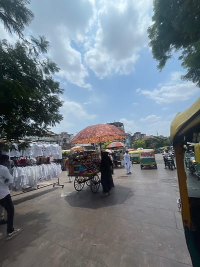 The image captures a street scene in Ahmedabad, India, under a bright, cloudy sky. In the foreground, a person in a white shirt and dark pants stands facing left, their arms crossed. To their right, a vendor pushes a cart filled with goods, sheltered by a large, patterned umbrella. The cart displays an assortment of items, possibly food or merchandise. Racks of white shirts hang to the left of the vendor, suggesting a clothing market. Further down the street, several auto-rickshaws are visible, some occupied by people, indicating bustling city life. Buildings with traditional Indian architecture line the background. The overall scene depicts a busy marketplace environment typical of Ahmedabad.