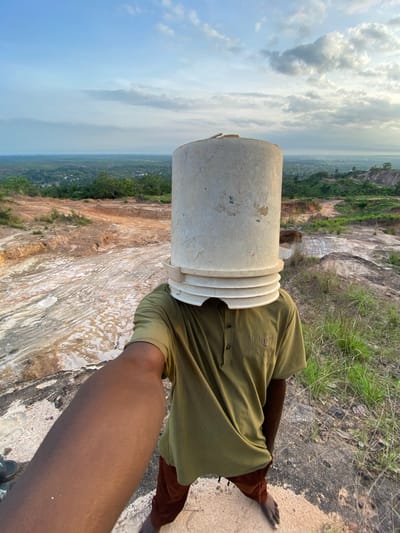 This image, originally rotated 90 degrees clockwise, depicts a person carrying a large, worn, off-white plastic bucket on their head, standing on the edge of a rugged landscape. The person is visible from the torso down, wearing a ribbed olive green short-sleeved shirt and dark reddish-brown pants. Their bare feet are placed on the sandy, rocky ground. A segment of their right arm and shoulder extends into the foreground from the bottom right.
The scene is set outdoors, likely at a high vantage point overlooking an expansive area in Ufueseni, Nigeria. To the immediate right of the person, the ground is covered with sparse green grass and loose soil. To their left, a vast open-pit quarry or mining site dominates the middle ground, characterized by exposed layers of various shades of brown, white, and grey earth, indicating significant excavation. Beyond the quarry, a distant panorama of lush green trees and scattered buildings of a settlement stretches towards the horizon. The sky above is a light blue, dotted with scattered white and grey clouds, suggesting daytime, possibly morning or late afternoon, with diffused lighting.
The activity portrays the person engaged in head-carrying, a common method of transport. No specific event is discernible beyond this act of carrying the bucket. There is no visible text in the image.