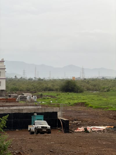 The image depicts a construction site in Vasai-Virar, India, on a cloudy day. In the foreground, a dirt area shows a white pickup truck with a green generator behind it. A partially constructed bridge or elevated structure runs across the middle ground. Beyond the bridge, a dense area of green vegetation and trees extends towards the horizon. In the background, a line of high-voltage electricity pylons are visible against a hazy mountain range under an overcast sky. The overall impression is one of ongoing development in a semi-rural or suburban setting.