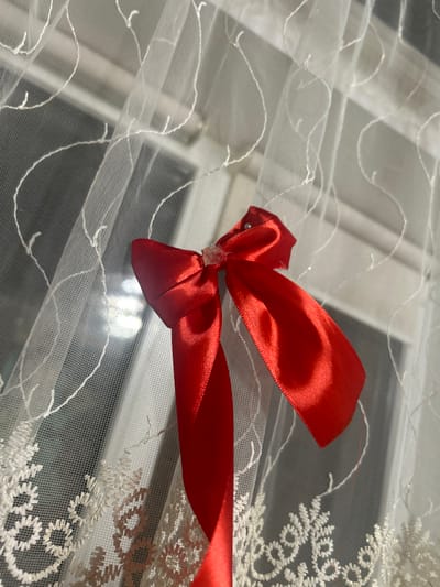 A red satin ribbon, tied in a bow with a clear central bead and a small white pearl accent on one loop, is positioned against a sheer white curtain. The curtain features two distinct white embroidered patterns: delicate wavy lines across its upper section and a denser, lace-like floral or geometric design along the bottom edge. Behind the translucent curtain, a window is visible, through which direct sunlight illuminates the scene. No individuals are present. This indoor environment is situated in Dmitriyevskaya, Russia.