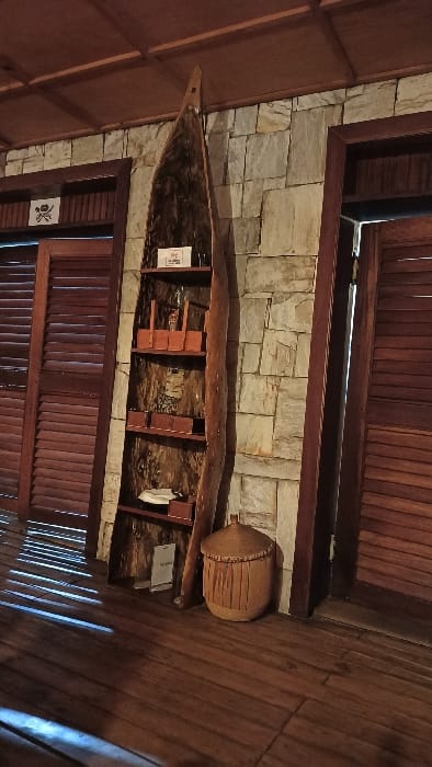A canoe-like wooden display, mounted on a light stone wall, is adorned with small wooden shelves and two informational plaques. A woven basket rests near the canoe’s stern. The surrounding architecture features dark wood paneling, creating a rustic, interior setting. The photo's orientation gives a unique perspective, as if viewing the scene from below, enhancing the unusual placement of the canoe.