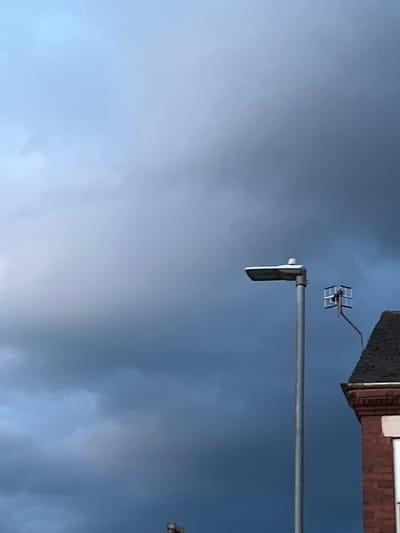 An exterior, eye-level, medium shot shows a grey sky filled with dark, ominous clouds. In the center-right of the frame, a tall, silver lamppost stands vertically. Attached to the top of the lamppost is a modern street light, angled to the left. To the right of the lamppost, a small, square aerial antenna, possibly for television reception, is mounted on a thin, orange rod. In the lower-right corner of the image, the dark, pitched roof of a red brick building is visible, along with a portion of its facade. The building appears to be a residential property. The image was captured in Stoke-on-Trent, United Kingdom.