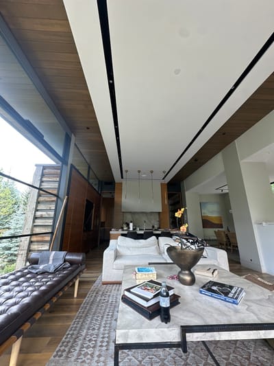 This image depicts a modern, high-end interior living space with an open-plan kitchen, located in Aspen, United States. No individuals are present in the scene.
The foreground features a rectangular coffee table with a light-colored, possibly stone, top positioned on a patterned area rug. On the table, a stack of books including one titled "OUR AMERICA" and "BOB ADELMAN," a single dark beverage bottle, a decorative dark metallic vase containing several yellow-orange orchids, and a small blue decorative box are arranged. Behind the coffee table is a large, light-colored, upholstered sofa draped with a black and white patterned throw. To the left, a dark brown tufted leather daybed with a grey and white striped blanket rests against a large expanse of floor-to-ceiling windows.
These windows reveal an exterior view of evergreen trees and a natural, possibly mountainous, landscape, along with a rustic wooden structure. The ceiling incorporates a multi-level design with a central white section bordered by dark wood paneling, featuring black linear track lighting and recessed circular lights.
In the mid-ground and background, an open-plan kitchen is visible, equipped with a dark-toned island, matching bar stools, light-colored countertops, and a sink. A modern range hood is centered above a cooktop area, and four slender, gold-topped pendant lights hang overhead. Integrated wood-paneled cabinetry is also present. Further to the right in the background, a dining area is partially visible, featuring a wooden table and chairs, with a large abstract painting displaying orange, yellow, and blue hues on the wall. The flooring throughout the space consists of light-toned hardwood.