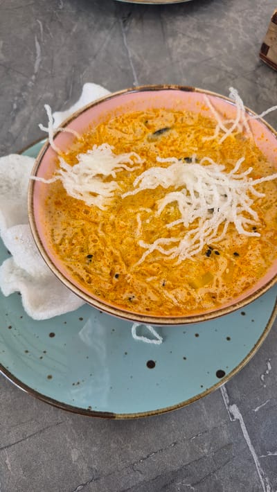 A close-up, overhead shot displays a bowl of soup with a creamy, orange broth and shredded toppings. The soup is garnished with white, crispy noodles and a few black seeds. To the left of the bowl are two pieces of white, puffed crackers. The bowl is a soft pink with a brown rim and sits on a light blue speckled plate. The plate rests on a dark, textured surface, likely a table. The background is a grey, marbled pattern. No people are visible in the image. The scene appears to be an indoor dining setting.