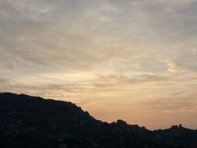 The image displays a landscape located in Hale Bandi Harlapura, India, under atmospheric conditions consistent with either sunrise or sunset. The upper two-thirds of the frame feature a sky filled with layered clouds, transitioning in color from light grey and white at the top to pale yellow, orange, and light peach tones near the horizon. The lower one-third of the image is occupied by a dark, silhouetted hilly terrain. This terrain is rugged, characterized by numerous prominent rocks and boulders, with indistinct sparse vegetation. No individuals, animals, man-made structures, or specific actions are discernible within the scene.