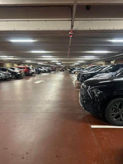 A long, narrow parking garage aisle stretching into the distance. Rows of parked cars line either side, with newer, dark-colored vehicles closer to the viewer and smaller, lighter ones further away. Overhead, fluorescent lights illuminate the aisle, reflecting off the concrete walls. A metal bar with red and green markers runs along the ceiling. The floor is a reddish-brown color.
