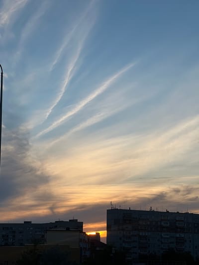 The image depicts a sunset over a city, likely in Stepnogorsk, Kazakhstan. The sky is filled with wispy clouds, transitioning from a pale blue on the left to vibrant shades of orange and yellow on the right, indicating the setting sun. The sun itself is partially visible, peeking through the clouds near the horizon. In the foreground, silhouetted against the bright sky, are the tops of several apartment buildings. These buildings are typical of Soviet-era architecture, with numerous windows and balconies. The overall scene evokes a sense of calm and the beauty of a natural phenomenon. The image is oriented vertically, with the buildings appearing to rise from the bottom of the frame. There is no visible text in the image.