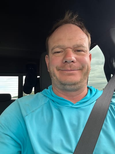 This media file features a portrait of a man in a vehicle. He appears to be in his 30s or 40s with light skin, brown hair, and a light beard. He is wearing a light blue long-sleeved shirt, and a grey seatbelt is visible across his chest. The man is looking at the camera with a slight smile, and his head is tilted. The background is blurred, but appears to be the interior of a car with a black dashboard and headrest. Through the window, a cloudy sky and what looks like buildings or trees are faintly visible. The lighting suggests it is daytime. The image is oriented sideways.