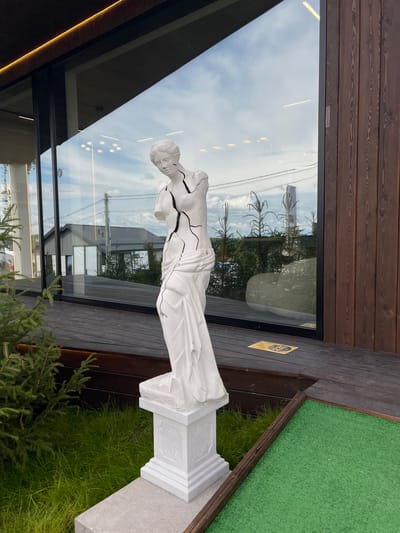A white, cracked statue of Venus de Milo stands on a white pedestal in front of a modern building with large windows and dark wood siding. The statue is partially broken, with visible cracks across its torso and arms. It is positioned on a wooden deck next to a patch of green grass and a small evergreen tree. In the reflection of the building's windows, a house and power lines are visible, suggesting an outdoor, possibly suburban or rural, setting. A small yellow sign with black text is on the wooden deck near the statue. The lighting suggests it is daytime, with soft clouds in the sky. The location is Yakunino, Russia.