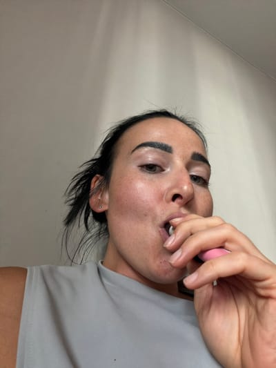A close-up, rotated image features a person, viewed from a low angle. The person has dark hair, dark eyebrows, and fair skin with freckles. They are wearing a light grey t-shirt. Their right hand is brought to their face, with fingers touching their mouth. A pink object, possibly a cosmetic item, is held between the fingers. The background is a plain, light-colored wall with a faint diagonal line, suggesting a corner or a seam. The lighting is soft, indicating an indoor setting, and the time of day is not discernible. The person's expression is neutral. No text is visible in the image.