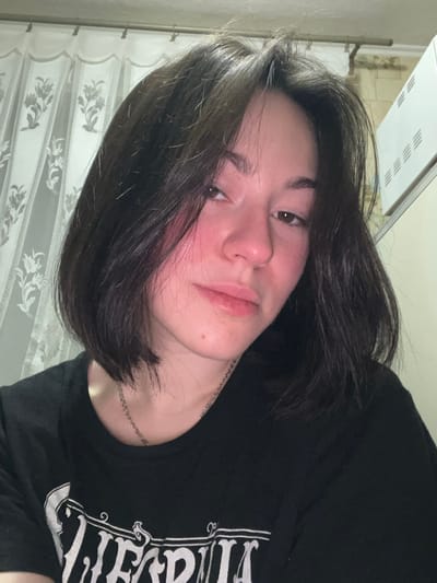 A young woman with dark, short hair, wearing a black graphic tee, is pictured indoors. Her face appears slightly flushed, and she wears a thin silver necklace. She has light skin and dark eyes. A window with patterned curtains is visible in the background, along with a bright, rectangular light fixture on the ceiling. The image is rotated to the right.