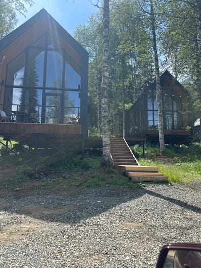 The image shows two modern, angular houses nestled amongst tall birch trees with lush green foliage. The houses feature large glass windows, reflecting the surrounding trees and the clear blue sky. One house is closer to the foreground, showcasing a multi-story design with wooden accents. A set of wooden stairs leads up to the entrance of one of the houses, built on a gravel path that extends to the right of the frame. The path is bordered by grass and more trees on the left. The scene appears to be set in a natural, possibly wooded environment, typical of a rural or vacation setting. The lighting suggests it is daytime, likely sunny given the bright sky and clear reflections. There is no visible text in the image. The overall impression is one of peaceful, contemporary architecture integrated into a natural landscape, potentially in Sheregesh, Russia, as indicated by the location context. The image is oriented vertically.