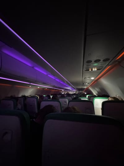 The image captures the dimly lit interior of an airplane cabin, viewed from a low angle looking down the aisle. The composition emphasizes the length of the cabin through strong leading lines created by the ambient lighting.

On the left side of the frame, overhead compartments feature a continuous strip of glowing purple LED light along their lower edge, casting a soft luminescence. Above this, a thinner orange LED strip runs higher along the cabin wall. Overhead service units, integrated into the cabin ceiling, are visible with small indicator lights, including a distinct green light on one panel and other yellowish-orange glows.

To the right, rows of airplane seats extend into the distance. Several passengers are visible, mostly from behind, their heads and shoulders appearing above the seatbacks. The seats have light-colored headrests and accents that appear to be a shade of green or teal, with the seat backs themselves showing darker hues, possibly purple or dark grey.

The overall setting suggests a night flight or a period when cabin lighting is intentionally dimmed, contributing to a tranquil or resting atmosphere. No specific activity beyond passengers being seated is discernible. There are no clear location cues within the image itself beyond it being an airplane cabin, and no legible text is visible.