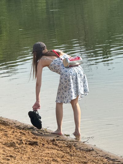 A person stands on a sandy shore near water. The person wears a floral dress, a baseball cap, and carries a black shoe in one hand and a metal container on their back. The water appears calm with tree reflections on the left side. The shore is sandy and uneven with some small plants or debris. The image suggests an outdoor setting by a lake or river. The location, Novosinkovo, Russia, isn't visually verifiable from the image.