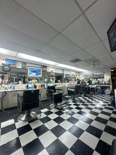 A barbershop interior features black and white checkered flooring and a tiled ceiling. Multiple barber chairs line the right side, with mirrors and framed pictures on the wall behind them. A customer is seated in one of the chairs, and a fan is visible in the background. The room has fluorescent lighting.