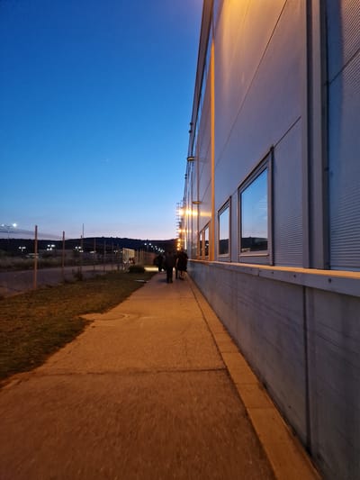 The image depicts an exterior view of a large, modern building with a corrugated metallic facade and rectangular windows, set against a twilight sky. A concrete walkway runs alongside the building, bordered by a strip of grass and a fence with multiple lights visible in the distance. Several individuals are walking along the path, silhouetted against the dim light. The scene appears to be in a semi-urban or industrial area, possibly on the outskirts of a city like Bahovitsa, Bulgaria, given the presence of modern infrastructure and the natural landscape in the background. The lighting suggests it is either dusk or dawn.