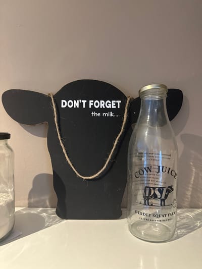 A black cow-shaped chalkboard with white lettering that reads "DON'T FORGET the milk...." hangs on a light-colored wall. A glass milk bottle with "COW JUICE" and "DIDDLY SQUAT FARM 1 LITRE PASTEURISED MILK" printed on it stands to the right of the chalkboard. The bottle is slightly to the right of center. A portion of a glass jar filled with white powder is visible on the far left. The items are displayed on a white surface. The scene is illuminated by artificial light, creating shadows on the wall and surface. The location is an indoor setting, likely a shop or home, in Oldemarkt, Netherlands.