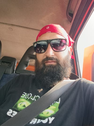 A man with a black beard and a red bandana is seen from the chest up, inside a vehicle. He is wearing black sunglasses and a black t-shirt with a green graphic and white text. A black seatbelt is across his chest. The interior of the vehicle, including the headrest and ceiling, are visible. The background is out of focus, but shows an orange interior panel of the vehicle and a bright sky. The image was taken at an unknown location in the city of Strumyani, Bulgaria.