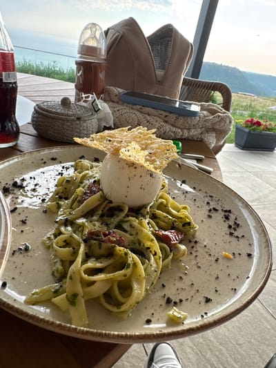 This media file captures a close-up of a plate of pasta served on a rustic wooden table. The pasta appears to be tagliatelle or fettuccine, tossed in a green sauce, possibly pesto, with visible chopped herbs and sun-dried tomatoes. A generous portion of burrata cheese sits atop the pasta, garnished with a delicate cheese crisp. The dish is sprinkled with what looks like black pepper or crushed olives, adding a touch of visual contrast.
In the background, blurred elements suggest an outdoor setting. A chair with a woven back and a knitted throw is visible to the left, along with a brown bag or backpack. Beyond this, a scenic view unfolds, with rolling hills and a hazy sky, indicative of a daytime scene, possibly late afternoon given the soft light. A glimpse of a balcony railing suggests the setting is elevated, perhaps a restaurant or cafe with a view.
To the left of the pasta, a baby bottle with a brown silicone holder is placed on the table, next to a small, woven decorative item. A partially visible bottle with a red label, possibly a drink, is in the foreground. The overall impression is a relaxed meal in a scenic location. The presence of the baby bottle hints at a family outing. No specific emotions are conveyed, but the scene suggests a moment of quiet enjoyment. The text on the bottle is too blurry to be legible. The location is identified as Kavarna, Bulgaria, which aligns with the hilly landscape.