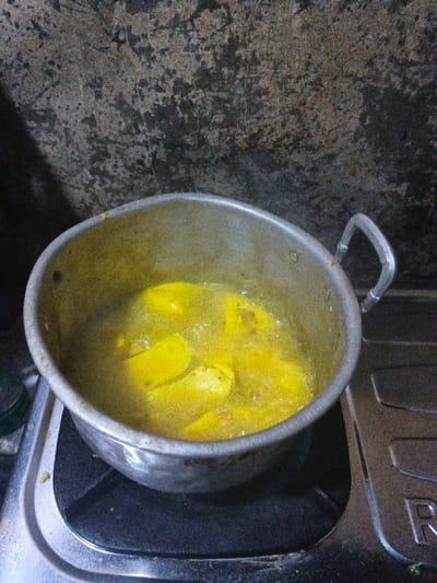 An overhead, slightly angled view captures a metal pot containing yellow food items boiling in a liquid, positioned on a gas stove burner. The pot is silver-colored, showing signs of use, with two handles visible. Inside, several irregularly shaped, bright yellow pieces of food are submerged in a bubbling, yellowish liquid. The pot sits on the left burner of a two-burner gas stove, which features a silver-toned metal surface and a black grate. A partial view of a brand logo, including the letter "R," is visible on the stove's right side. The background consists of a dark, textured wall, possibly concrete or painted plaster, exhibiting areas of wear and discoloration. The lighting is dim. This cooking activity takes place in a kitchen environment within thành phố Biên Hòa, Vietnam. No individuals are present in the frame.
