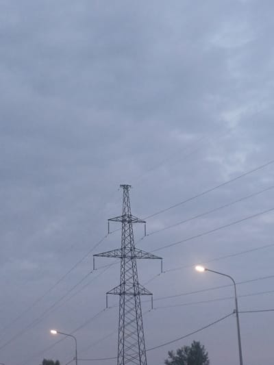 The image depicts an overcast sky with a tall, lattice-style power transmission tower dominating the center. Several parallel power lines crisscross the frame, extending from the tower. In the lower right portion of the image, a street lamp with a lit bulb is visible, mounted on a pole. A hint of greenery, likely trees, can be seen at the bottom edge of the frame. The scene appears to be an outdoor urban or semi-urban environment. The location provided is Elitnyi, Russia.