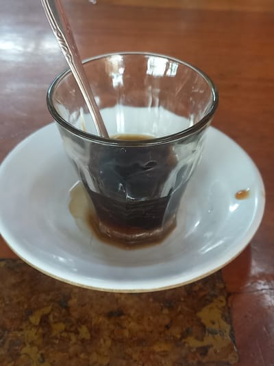 A close-up, high-angle shot shows a glass of dark coffee on a white saucer. The glass is partially filled with coffee, and a silver spoon rests inside. The saucer is positioned on a dark, wooden table. The table surface appears worn and has a textured pattern. The background is blurred, suggesting a shallow depth of field. The image is well-lit, with reflections visible on the glass and saucer. The scene is devoid of any people or animals.