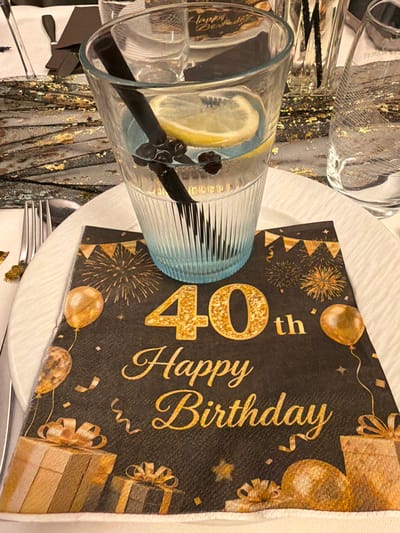 This is a close-up image of a festive table setting, capturing details of a celebration. The overall theme appears to be black and gold, conveying an elegant and celebratory atmosphere.
At the center of the frame is a clear glass tumbler with a subtle light blue tint and a ribbed or fluted texture. Inside the glass, a beverage contains a bright yellow lemon slice and several dark, small berries, possibly blueberries or blackcurrants. A dark-colored stir stick or straw is also present in the drink.
Beneath the glass, a white plate is visible, and to its right, a black napkin is prominently displayed. The napkin features glittery gold text, clearly stating "40th Happy Birthday" in a decorative font. The napkin's design is further enhanced with gold illustrations of balloons, bursting fireworks, scattered confetti, and wrapped gift boxes against the black background, strongly indicating a 40th birthday event.
To the left and slightly behind the central glass, parts of another clear glass are visible, along with decorative elements that include black fabric or ribbon with shimmering gold flecks, suggesting a table runner or other elaborate decor. Silver cutlery can be partially seen near the edge of the white plate.
The image depicts an indoor setting, focusing solely on the table arrangement, with no people visible. The specific location context of "Grass, Luxembourg" is not supported by any visual cues within this indoor close-up shot. The scene suggests a sophisticated and joyful adult birthday celebration.