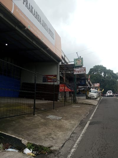 The image displays a commercial street scene under an overcast sky. On the left, a large industrial-style building features a prominent white and orange sign reading "PRAKARSA JAYA STEEL," with the address "Jl Cigombong Baros No 5 Rt03 / Rw01 Ds. Cigombong Kec. Ciawi Bogor 41725, Indonesia" and two phone numbers. The building's open entrance, dark inside, is secured by a black metal grid fence, behind which stacked rectangular materials and a red banner are visible.
Adjacent to this, further down the street, another business operates under a metal awning. A green sign above it shows a lion logo and "SBN PRAKARSA LIMA STEEL," with a secondary sign below stating "SUPPLIER BESI & BAJA" (Supplier of Iron & Steel). A white passenger van is parked on the concrete curb directly in front of this establishment.
The foreground consists of a worn concrete sidewalk with a rectangular metal drainage grate. At the far left base of the curb, a small patch of weeds and scattered litter, including plastic bags and paper, is present. An asphalt road, marked by a faded white line, runs along the right side of the frame. Further down the road, two additional white vehicles, a van and an SUV, are visible in traffic. The background on the right is filled with dense green foliage from large trees. No discernible individuals or specific actions are visible within the scene.