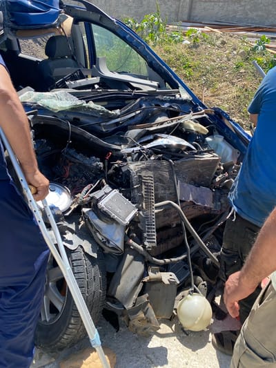 The image depicts a severely damaged blue car, possibly after an accident, located in what appears to be an outdoor area with dirt and some vegetation, potentially near a residential or industrial area in Baraki, Algeria. The car's front end is heavily crushed, exposing the engine components, radiator, and other internal parts. Shattered glass is visible. Two individuals are present: one wearing a blue shirt is holding a crutch, and another individual is partially visible, wearing a gray shirt. The presence of the crutch suggests possible injury to one of the individuals. The focus is on the wrecked vehicle, suggesting a situation involving damage or loss.