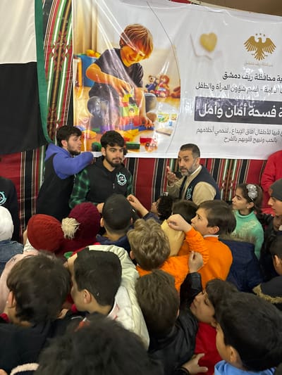 This image captures an indoor community event in Kafr Batna, Syria, featuring a large group of children interacting with several adults. In the background, a prominent white banner displays an image of a smiling child playing with colorful building blocks. Arabic text on the banner identifies it with the "Damascus Countryside Governorate," "Women and Child Affairs Department," and states "A Space of Safety and Hope," accompanied by an eagle emblem. Behind and to the sides of the banner, a patterned fabric, resembling traditional textiles in shades of brown, black, white, and green, is visible, alongside a partial view of the Syrian flag on the far left.
In the mid-ground, an adult male with a beard, wearing a light brown vest over a patterned shirt, is actively speaking and gesturing with his right hand towards the assembled children. To his immediate left stand two younger adult males; one with a beard, wearing a dark vest over a green plaid shirt with a logo, and another in a blue hoodie over a dark top, looking towards the speaking adult. They appear to be staff or volunteers.
The foreground and mid-ground are densely populated by approximately two dozen children, predominantly young boys and some girls, all facing towards the adults. They are dressed in various casual winter clothing, including jackets, sweaters, and knitted hats, such as a child with a red hat and a white pom-pom. Several children are attentive to the speaking adult, with some raising their hands or reaching forward, indicating an interactive or distribution event. A girl on the far right, wearing a light green sweater and a sparkling hair accessory, observes the scene. The overall scene depicts an organized gathering for children in what appears to be a community setting.