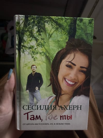 A close-up image displays a person holding a paperback book titled "Там, где ты" (There, where you are) by "СЕСИЛИЯ АХЕРН" (Cecelia Ahern). The text further identifies her as "ОТ АВТОРА БЕСТСЕЛЛЕРА «P.S. Я ЛЮБЛЮ ТЕБЯ»" (From the author of the bestseller "P.S. I Love You").

The book cover features a vibrant green background, depicting trees and foliage, suggesting a natural, possibly wooded or park-like setting. In the upper right portion of the cover, a woman with dark hair is prominently featured, smiling broadly towards the viewer. In the lower-middle left, a man with light brown hair is shown standing, dressed in a dark jacket and light pants, also looking towards the viewer amidst the green backdrop.

A hand, with light blue nail polish, holds the bottom right edge of the book, partially visible in the lower right corner of the photograph. The background behind the book and hand is dark and out of focus, indicating an indoor setting or low light conditions. The book is held at a slight angle.
