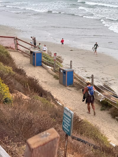 The image depicts a sandy beach with an overcast sky. Several people are present on the beach and walking along a sandy path that leads down to the beach. One person, a surfer with a surfboard, is walking up the path away from the ocean. Another person is walking down the path with a surfboard and a backpack. Two blue trash and recycling bins are situated along the path. A warning sign is posted near the path. The environment includes dry brush and vegetation along the sides of the path and sand dunes. The ocean waves are visible in the background. The scene appears to be located in Encinitas, California, United States of America, given the signage and typical beach environment.